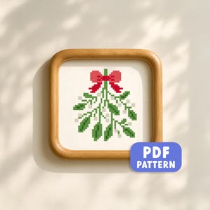 Puede incluir: Un patrón de punto de cruz enmarcado de muérdago con un lazo rojo. El diseño presenta hojas verdes y bayas blancas sobre un fondo crema. El marco es de madera marrón claro, y una etiqueta azul dice "PDF PATTERN".