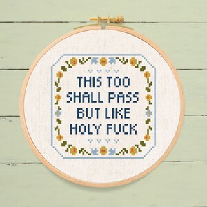 May include: Cross-stitch embroidery in a wooden hoop. The design features the text "THIS TOO SHALL PASS BUT LIKE HOLY FUCK" in blue, surrounded by a floral border with yellow and blue accents. The background is a light beige fabric.