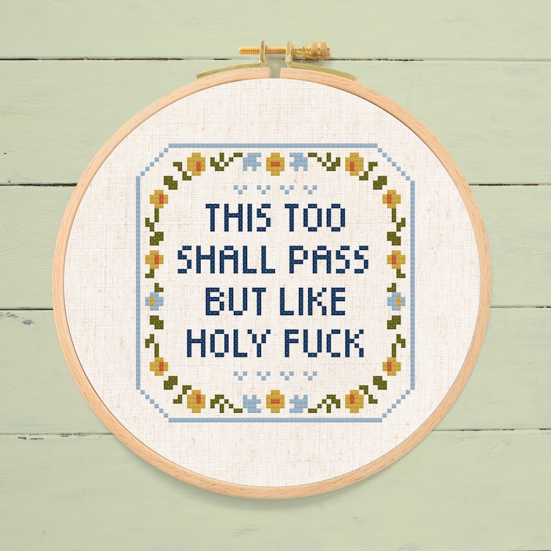 May include: Cross-stitch embroidery in a wooden hoop. The design features the text "THIS TOO SHALL PASS BUT LIKE HOLY FUCK" in blue, surrounded by a floral border with yellow and blue accents. The background is a light beige fabric.