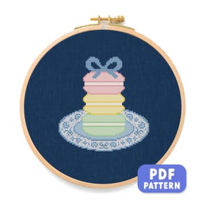 May include: Cross-stitch embroidery of a stack of colorful macarons with a bow on top, displayed on a decorative plate. The design is set against a dark blue fabric within a wooden embroidery hoop. The text "PDF PATTERN" is in a blue box.