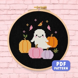 以下が含まれることがあります： 黒い背景に、笑顔の白い幽霊とオレンジ、ピンク、紫色のカボチャが描かれたクロスステッチパターン。木製の刺繍枠に額装されています。「PDF PATTERN」というテキストが青いボックスに入っています。