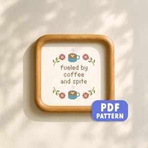 May include: Cross-stitch art in a wooden frame. The design features the text "fueled by coffee and spite" with coffee cups and floral accents. A blue PDF pattern button is in the lower right corner.