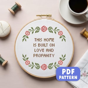 May include: A cross-stitch embroidery hoop with the text "THIS HOME IS BUILT ON LOVE AND PROFANITY" surrounded by a floral border. The design features pink roses and green leaves. A PDF PATTERN button is in the lower right corner.
