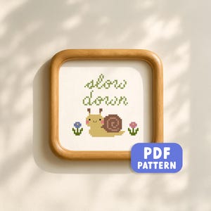 May include: Cross-stitch pattern framed in a light wood. The design features a smiling snail with the words "slow down" in green. Small blue and pink flowers flank the snail. A blue "PDF PATTERN" button is in the lower right corner.