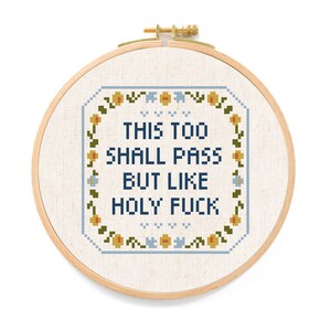 May include: Cross-stitch embroidery in a wooden hoop. The design features the text "THIS TOO SHALL PASS BUT LIKE HOLY FUCK" in dark blue, surrounded by a floral border with yellow, blue, and green accents. The background is a light beige fabric.