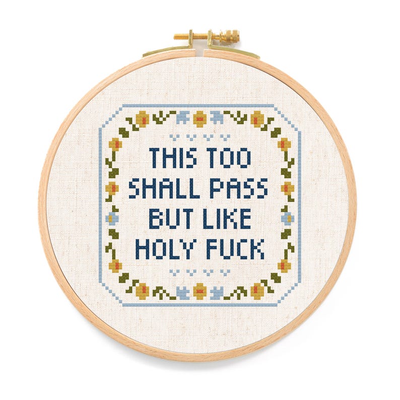 May include: Cross-stitch embroidery in a wooden hoop. The design features the text "THIS TOO SHALL PASS BUT LIKE HOLY FUCK" in dark blue, surrounded by a floral border with yellow, blue, and green accents. The background is a light beige fabric.