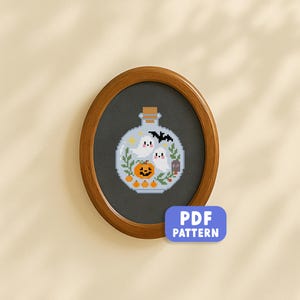 May include: A framed cross-stitch design depicting a Halloween scene within a glass bottle. The design includes two ghosts, a pumpkin, a bat, and the text "RIP". The frame is oval and wooden. The image also includes a "PDF PATTERN" label.