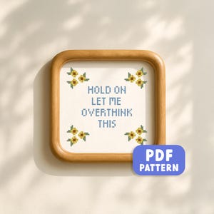 May include: A framed cross-stitch with the text "HOLD ON LET ME OVERTHINK THIS" in blue pixelated letters. The design includes yellow and green floral accents. A blue rectangle with "PDF PATTERN" is in the lower right corner.
