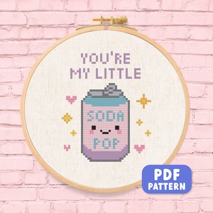 May include: A cross-stitch design in a wooden hoop. The design features the text "YOU'RE MY LITTLE" above a cartoon soda can with the words "SODA POP". Pink hearts and gold stars surround the soda can. A blue rectangle with "PDF PATTERN" is in the bottom right.