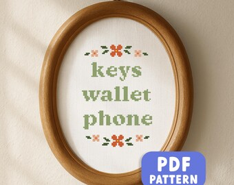 May include: Oval wooden framed cross-stitch with the words "keys wallet phone" in green. The cross-stitch has floral accents in pink and orange. A blue PDF pattern button is in the lower right corner.