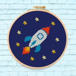 Op de afbeelding: Een blauw en wit raket met oranje vlammen die door een donkerblauwe ruimte vliegt met gele sterren. De raket is geborduurd op een ronde houten ring.