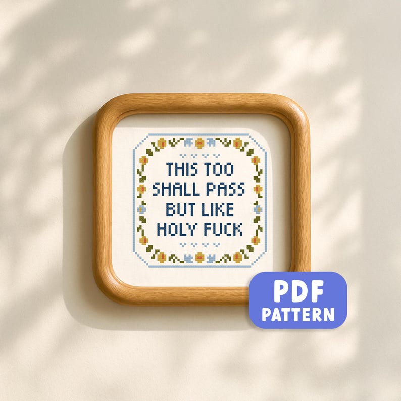 May include: A framed cross-stitch with the text "THIS TOO SHALL PASS BUT LIKE HOLY FUCK" in a square wooden frame. The cross-stitch has a floral border in blue, yellow, and green. A blue PDF pattern button is in the lower right corner.