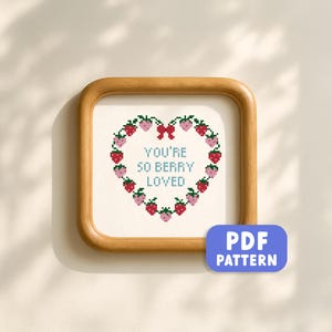 May include: Cross-stitch art in a wooden frame. The design features a heart-shaped wreath of red and pink strawberries with a red bow at the top. The words "YOU'RE SO BERRY LOVED" are in the center. A blue "PDF PATTERN" button is in the lower right.