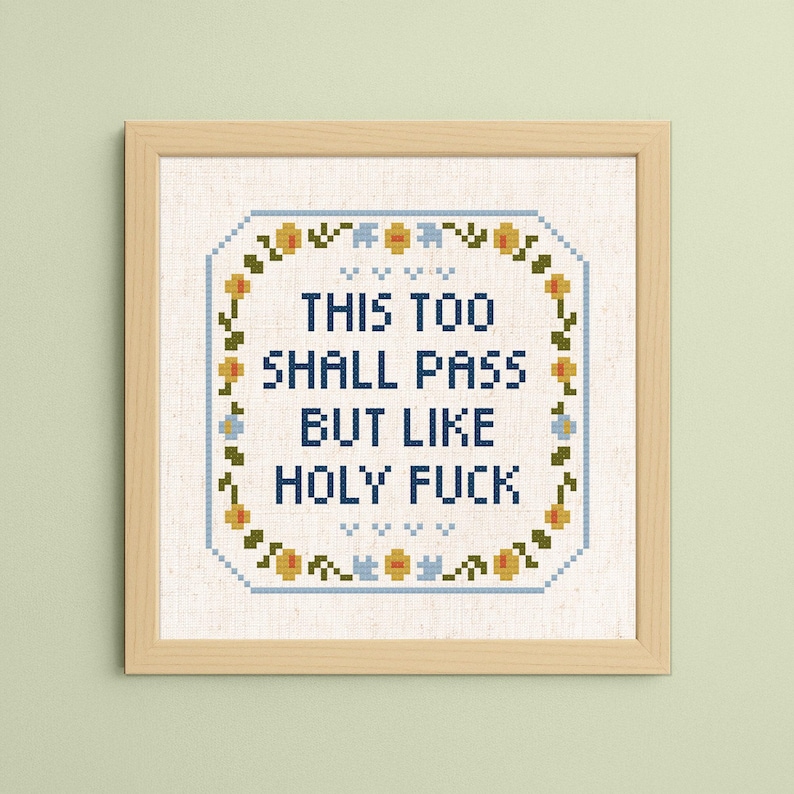 May include: Cross-stitch art in a wooden frame. The text reads "THIS TOO SHALL PASS BUT LIKE HOLY FUCK" in blue letters. The design is surrounded by a floral border with yellow and blue accents.