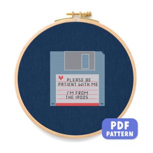 Puede incluir: Patrón de punto de cruz sobre tela azul, en un aro de madera. El diseño presenta un disquete pixelado con el texto "PLEASE BE PATIENT WITH ME, I'M FROM THE 1900S". Un patrón PDF está disponible.