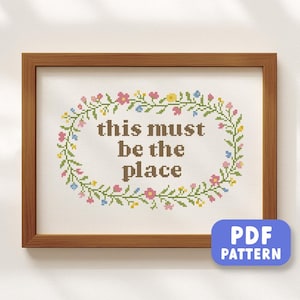 Puede incluir: Una obra de arte de punto de cruz enmarcada con el texto "this must be the place" en marrón. El texto está rodeado por una corona floral en rosa, azul y amarillo. Un botón azul "PDF PATTERN" está en la esquina inferior derecha.