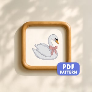 May include: A framed cross-stitch pattern of a white swan with a pink bow. The swan has a black eye and an orange beak. The framed artwork is square with rounded corners and a light wood finish. A blue rectangle with the text "PDF PATTERN" is in the lower right corner.