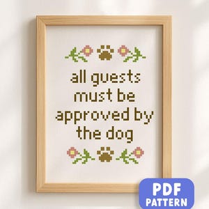 Puede incluir: Obra de arte en punto de cruz enmarcada con el texto "all guests must be approved by the dog". El diseño incluye huellas de patas y detalles florales. El marco es de color marrón claro y el fondo es blanquecino.