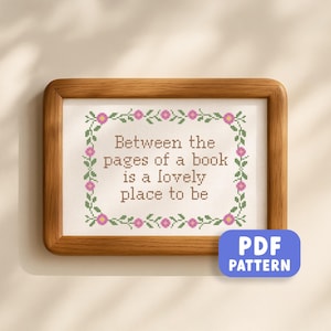 May include: A wooden framed cross-stitch sampler with the quote "Between the pages of a book is a lovely place to be." The design includes a floral border with pink flowers and green leaves. A blue "PDF PATTERN" button is visible.