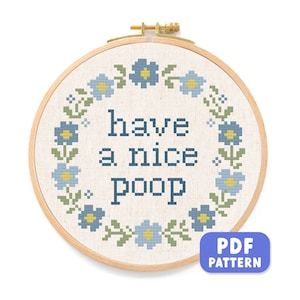 Puede incluir: Un bastidor de bordado de punto de cruz redondo con el texto "have a nice poop" en azul oscuro. El diseño incluye un borde floral con flores azules y amarillas y hojas verdes. También hay un patrón PDF disponible.