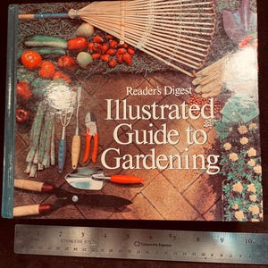May include: A hardcover book titled "Reader's Digest Illustrated Guide to Gardening" with a colorful cover featuring gardening tools and produce. A stainless steel ruler is placed below the book. The book's cover displays a rake, tomatoes, and other gardening items.