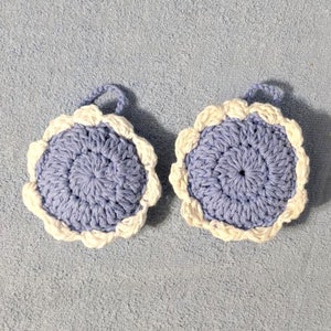 May include: Two crocheted lavender-colored scrubbers with white edging. The scrubbers are round and have a textured surface.