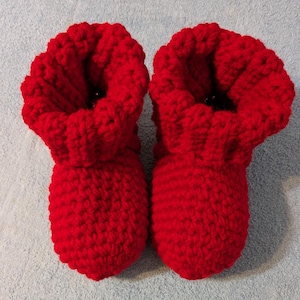 May include: A pair of bright red, handmade crochet baby booties. The booties have a textured, chunky knit appearance with a folded-over cuff. The booties are designed to keep a baby's feet warm and cozy.