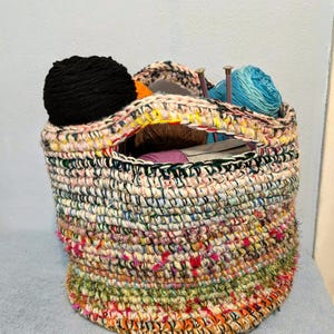 May include: A handmade, cylindrical crochet basket filled with yarn balls and knitting needles. The basket features a colorful, textured design with a mix of pink, green, orange, and black threads. The open top allows easy access to the contents.