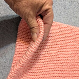 May include: Close-up of a knitted item in a soft coral color. The texture of the knit is visible, with the stitches creating a ribbed effect along the edge. The item is being held, showing the detail of the yarn.