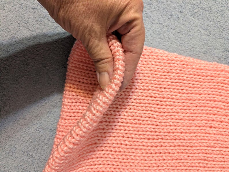 May include: Close-up of a knitted item in a soft coral color. The texture of the knit is visible, with the stitches creating a ribbed effect along the edge. The item is being held, showing the detail of the yarn.