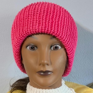 May include: A vibrant pink knitted beanie hat. The hat is made of a ribbed knit fabric and is displayed on a mannequin head. A white crocheted collar with a scalloped edge is also visible.