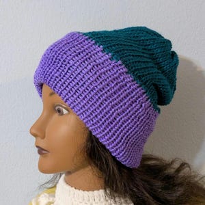 May include: A two-toned knit beanie in shades of purple and teal. The hat is made of a ribbed knit fabric and has a folded brim. The beanie is displayed on a mannequin head. This hat is a stylish accessory for cold weather.