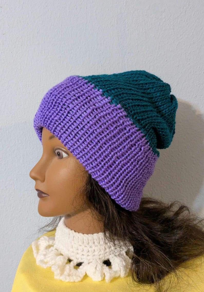 May include: A two-toned knit beanie in shades of purple and teal. The hat is made of a ribbed knit fabric and has a folded brim. The beanie is displayed on a mannequin head. This hat is a stylish accessory for cold weather.