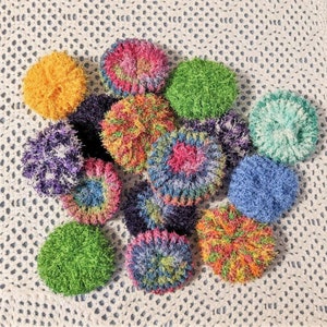 Puede incluir: Una colección de paños de cocina de ganchillo de colores en varios patrones y colores, incluyendo verde, azul, morado, rosa y amarillo. Los paños de cocina están dispuestos en una pila sobre una superficie de ganchillo blanco.