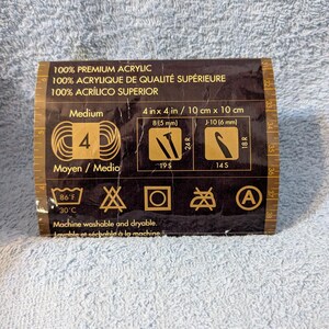 Puede incluir: Una etiqueta marr&oacute;n con texto y s&iacute;mbolos para hilo. La etiqueta dice "100% Premium Acrylic" e incluye medidas de 10 cm x 10 cm. Tambi&eacute;n muestra instrucciones de lavado y tama&ntilde;os de agujas.