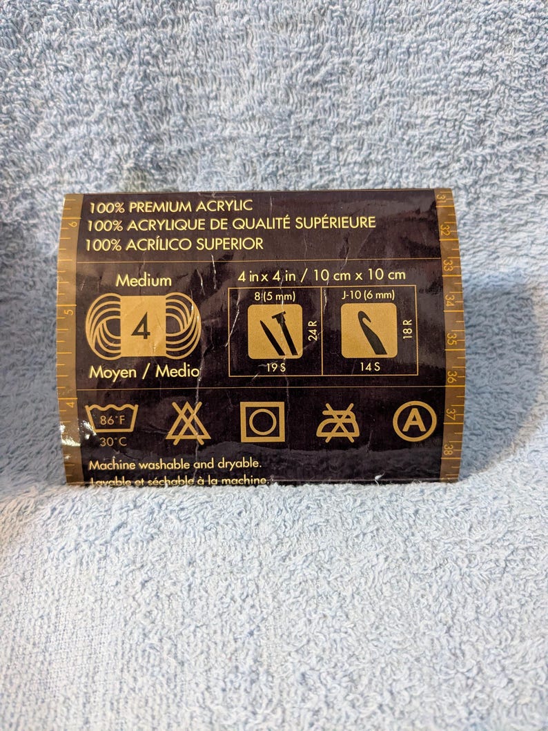 Puede incluir: Una etiqueta marr&oacute;n con texto y s&iacute;mbolos para hilo. La etiqueta dice "100% Premium Acrylic" e incluye medidas de 10 cm x 10 cm. Tambi&eacute;n muestra instrucciones de lavado y tama&ntilde;os de agujas.