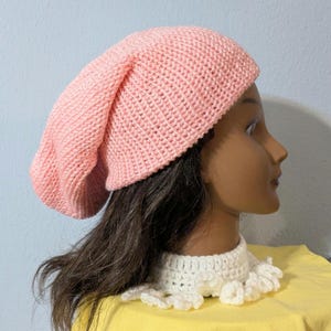 May include: A light pink knitted beanie hat. The hat is made of a soft, textured yarn and has a slouchy fit. The hat is shown on a mannequin head with dark brown hair and a white crocheted collar.