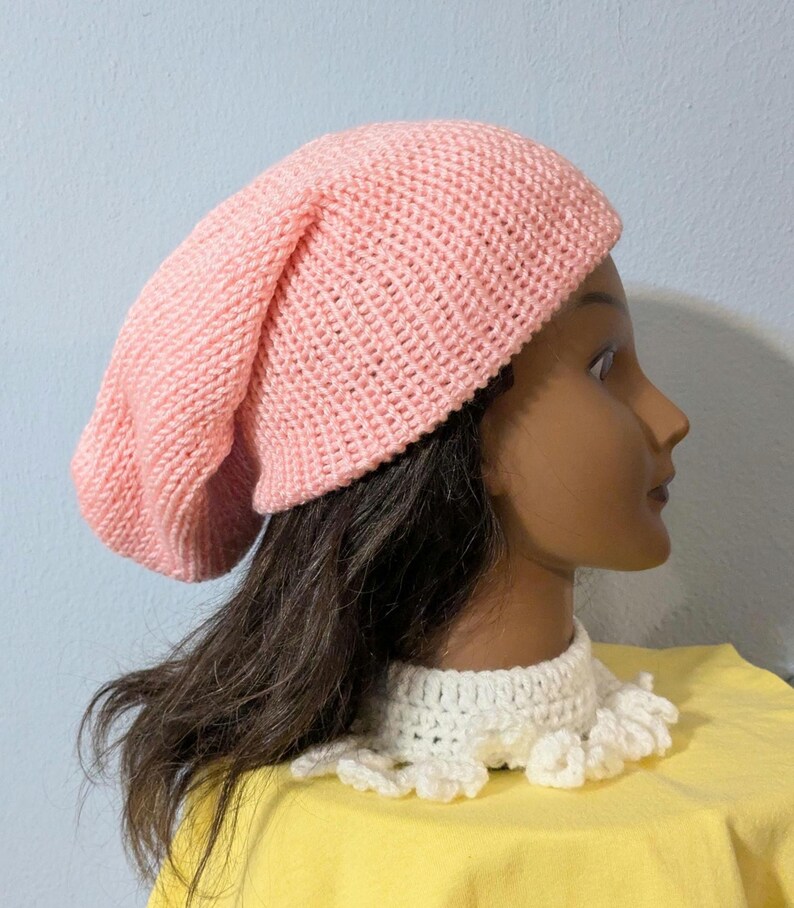 May include: A light pink knitted beanie hat. The hat is made of a soft, textured yarn and has a slouchy fit. The hat is shown on a mannequin head with dark brown hair and a white crocheted collar.