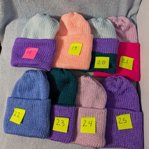 May include: A collection of eight knitted beanies in various colors, including blue, pink, purple, and green. Each beanie has a folded cuff and a small yellow tag with a number on it. The beanies are arranged on a light blue surface.
