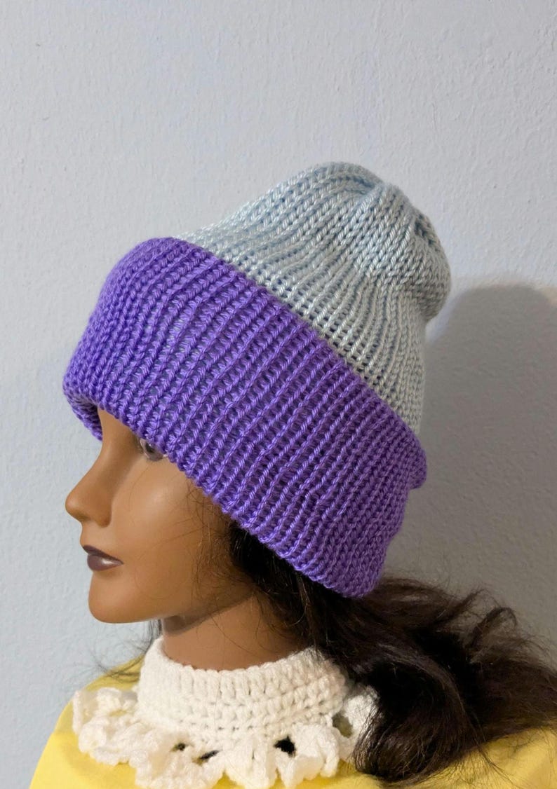 May include: A two-tone knit beanie in a light blue and purple color scheme. The hat features a ribbed knit design, with the purple section forming the brim. The beanie is displayed on a mannequin head.