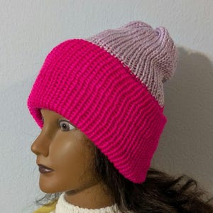 May include: A two-tone knitted beanie hat. The lower portion is a vibrant fuchsia pink, while the upper section is a soft lavender. The hat is made of a thick, textured yarn, suggesting warmth and comfort. The hat is a winter accessory.