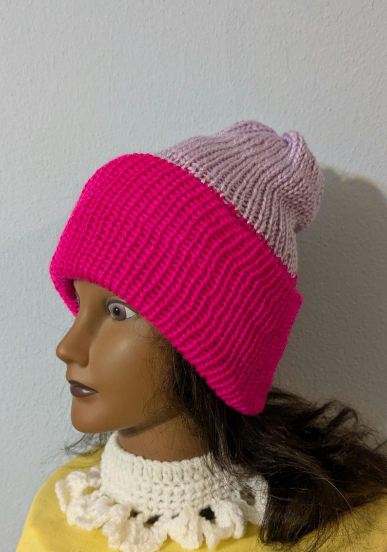 May include: A two-tone knitted beanie hat. The lower portion is a vibrant fuchsia pink, while the upper section is a soft lavender. The hat is made of a thick, textured yarn, suggesting warmth and comfort. The hat is a winter accessory.