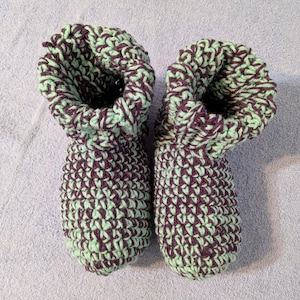 May include: A pair of handmade crochet booties in a two-tone pattern. The booties are primarily mint green with a dark purple accent color. The tops are folded over, revealing the interior. The booties are resting on a light blue surface.