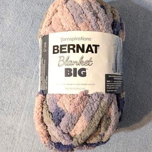 May include: A large, soft ball of Bernat Blanket Big yarn in shades of pink, purple, and gray. The yarn is labeled with the brand name and product details. Ideal for creating cozy blankets and other craft projects.