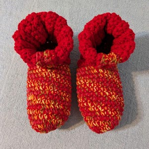 May include: A pair of handmade crochet slippers. The slippers are primarily red with a textured stitch pattern. The cuffs are a solid red, while the foot area features a mix of red, yellow, and orange yarns. The slippers are designed to keep feet warm.