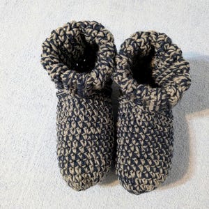May include: A pair of handmade crochet slippers in a two-tone pattern of navy blue and beige. The slippers have a bootie-style design with a folded-over cuff. The thick yarn creates a textured appearance, perfect for cozy indoor wear.