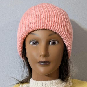 May include: A light pink knitted beanie hat. The hat is made of a ribbed knit fabric. The hat is on a mannequin head. The mannequin is wearing a white crocheted collar and a yellow shirt.