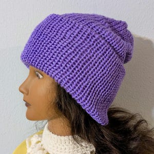 May include: A solid purple knit beanie hat. The hat is made of a ribbed knit fabric and has a folded brim. The hat is shown on a mannequin head with dark brown hair and a white lace collar.