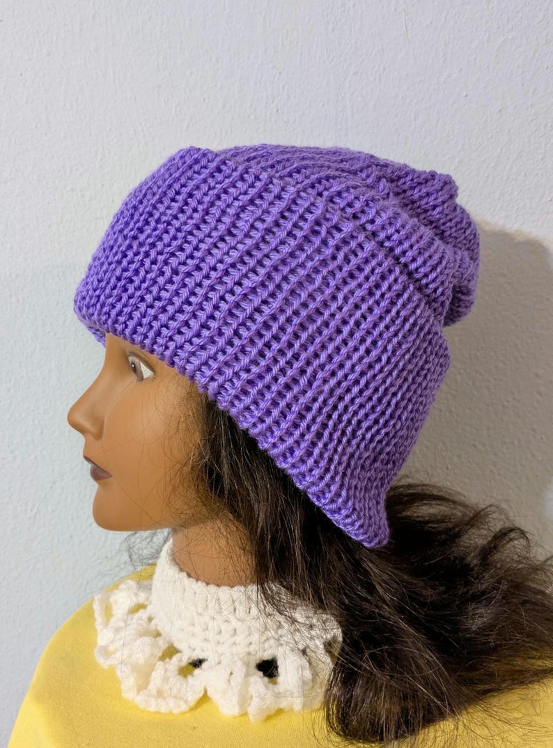 May include: A solid purple knit beanie hat. The hat is made of a ribbed knit fabric and has a folded brim. The hat is shown on a mannequin head with dark brown hair and a white lace collar.