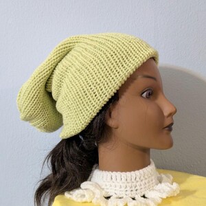 Puede incluir: Un gorro tejido de color verde claro. El gorro est&aacute; hecho de una tela de punto acanalado y tiene un ajuste holgado. El gorro se muestra en una cabeza de maniqu&iacute; con cabello oscuro y un cuello de ganchillo blanco.